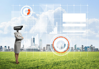 Camera headed woman standing on green grass against modern cityscape