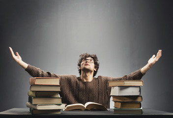 Helpless young man sitting at the desk full of books.