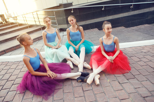 Ballet dancers sitting and talking on street. Young ballerinas in color tutus sit on red brick stone floor. Ballet feet on the point