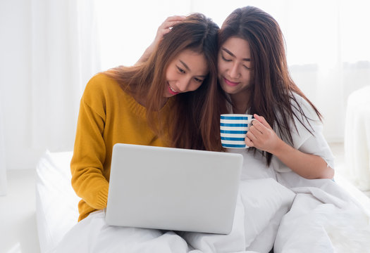 Asia Lesbian Lgbt Couple Sitting On Bed Hug And Using Laptop Computer While Drinking Hot Coffee Cup And Near White Window Sunlight In Bedroom With Happiness Moment Together,love Wins Concept.