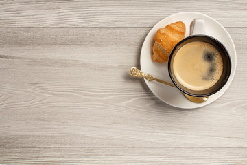 Top view of cup of black coffee with fresh croissant and spoon on saucer with copy space