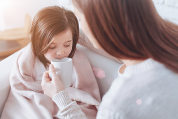Tasty tea. Attractive brunette holding white cup in left hand and looking at her daughter while sitting in front of her
