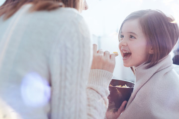 Express positivity. Little female person sitting in semi position while looking at her mother that feeding her
