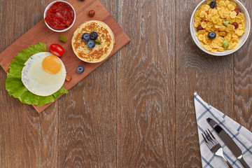top view of pancake with blueberries and fried eggs with tomatoes and lettuce on a desk and a cornflakes in a bowl, with empty space