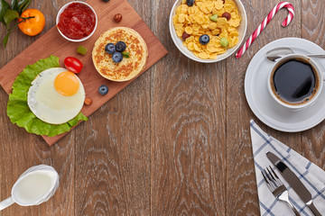 top view of pancake with blueberries and fried eggs with tomatoes and lettuce on a desk and a cornflakes in a bowl, and a cup of coffee with empty space