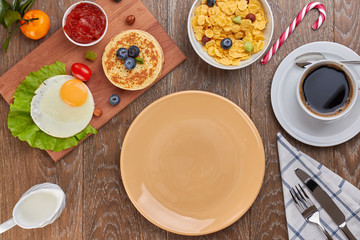 top view of empty brown plate,  pancake with blueberries and fried eggs with tomatoes and lettuce on a desk and a cornflakes in a bowl, and a cup of coffee