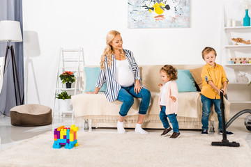 smiling pregnant woman looking at adorable little kids cleaning room with vacuum cleaner at home