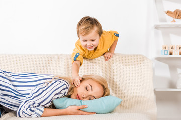 smiling little boy touching mother sleeping on sofa at home