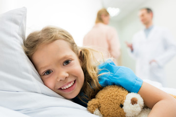 happy kid lying on hospital bed while mother talking with doctor