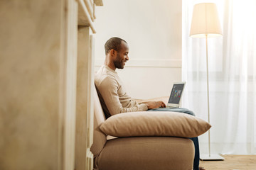 Working day. Good-looking young inspired afro-american businessman smiling and working on his laptop while sitting on the sofa