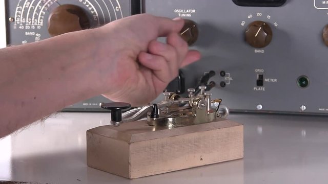 Hand makes equipment adjustments and sends morse code message on telegraph key for radio transmission with vintage electronic hardware engineering military lab test equipment device in the background