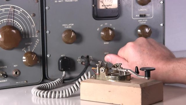 Telegraph operator makes equipment adjustments and sends morse code radio transmission with vintage electronic hardware engineering military lab test equipment device in the background