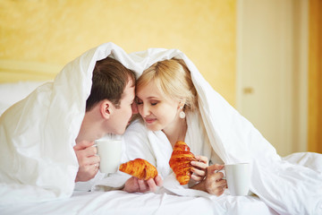 Couple in white bathrobes in bed, eating croissants