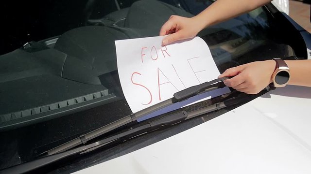 Closeup slow motion footage of young woman putting for sale sign under car windshield wipers. Perfect footage for selling automobile