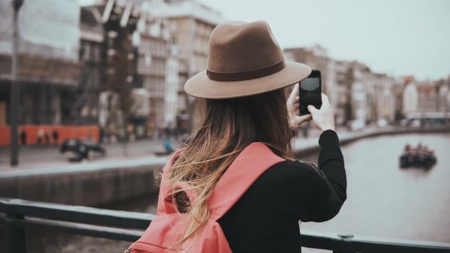 Young girl in hat takes photos of scenic river. Girl with long hair and red backpack sharing pictures on her blog. 4K.