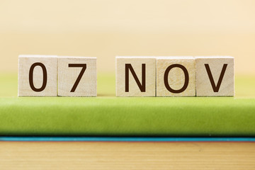 Wooden cube shape calendar for NOV 7 on green book, table.