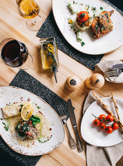 a beautiful serving of two dishes roasted quail with mashed potatoes and mushrooms and baked meat with a gratin with a glass of wine, a cherry branch and a bottle of olive oil on a wooden table