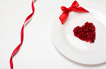 Pomegranate grains in a heart shape on white plate and red ribbon.On white background.Background for valentine's day.Minimalism.Copy space.Closeup