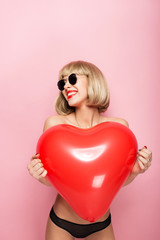 Sexy blonde hugging in the shape of a heart a red ball and smiling. On a pink background.