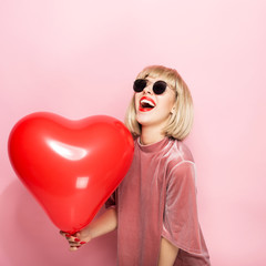 Sexy blonde hugging in the shape of a heart a red ball and smiling. On a pink background.