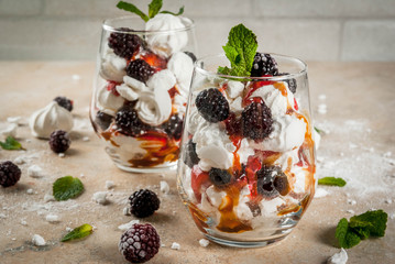 Traditional English dessert. Eton mess - whipped cream, meringue, fresh blackberries, sauce and caramel. In serving glasses on a light stone table. Copy space