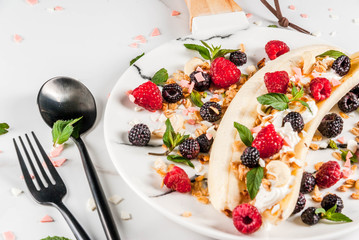 Summer fruit berry breakfast. Healthy banana split breakfast with cream cheese, raspberries, blackberries, mint, white and pink chocolate. On white marble plate, marble table. Copy space