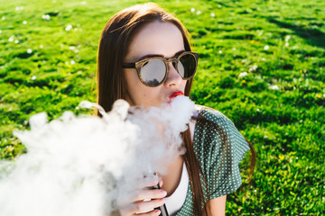 Happy fashion smiling Woman in sunglasses smoking vape on street,smoke
