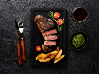 Meat Picanha steak, traditional Brazilian cut with potato wedges, chimichurri sauce and rosemary on black meat cutting board. Steak and a glass of red wine with fork and knife.