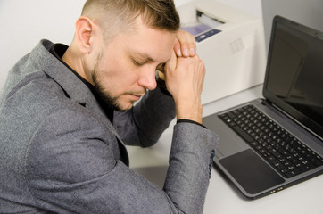 tired man with closed eyes and head leaning against hands at table with laptop in office