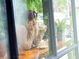 Beautiful Asian woman wearing white sweater and drinking a cup of coffee in glass house cafe, alone, single concept