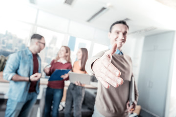 Shake my hand. Handsome joyful pleasant man standing in the room near his colleagues stretching out his hand and feeling good himself.