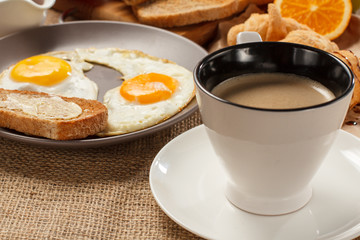 Cup of black coffee, plate with fried eggs and toast with butter, croissants, bread and fresh orange