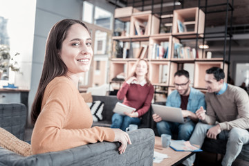 Trust my colleagues. Smiling pretty confident woman sitting in the room on the sofa turning her head to the right and smiling.