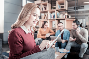 Useful gadget. Joyful occupied smart woman sitting in the room near her friends smiling and using the tablet.