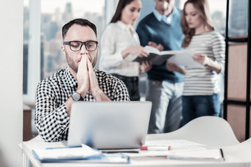 Must to do this. Serious concentrated thoughtful man sitting in the room by the table near his colleagues holding hands near lips and working with the laptop.