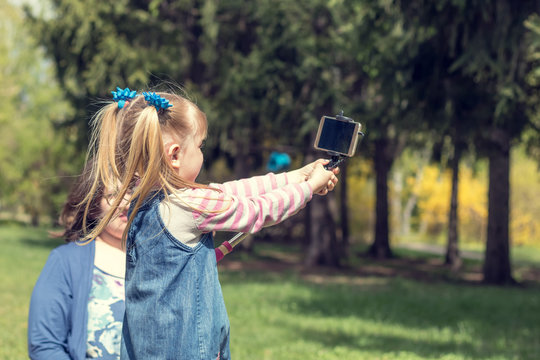 Cute little blonde girl with two ponytails taking selfie in the city park on a spring sunny day