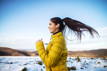 Young active athletic girl jogging in winter sportswear on snowy winter road with earphones in the sunny morning.