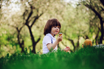 handsome stylish 3 years old toddler child boy with funny face in suspenders enjoying sweets on picnic in spring or summer garden or park and feeding his teddy bear