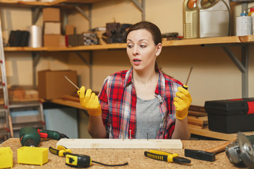 Upset perplexed caucasian young woman in plaid shirt, gray T-shirt, yellow gloves twisting by screwdriver screw, working in carpentry workshop at wooden table place with piece of wood, different tools