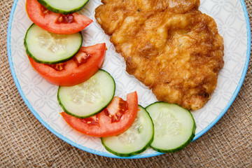 Plate with fried meat chop in egg batter and sliced tomato and cucumber