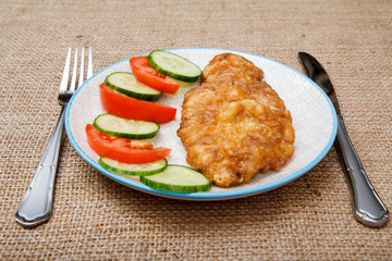 Plate with fried meat chop in egg batter and sliced tomato and cucumber with knife and fork
