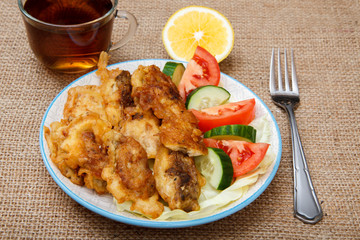 Plate with pieces of fried fish and freshly sliced tomatoes and cucumbers, cup of black tea with slice of lemon