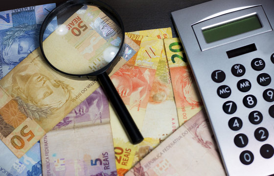 Notas de dinheiro acompanhado de moedas, carteira de trabalho, lupa, calculadora encima da mesa