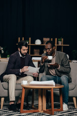 multiethnic young businessmen sitting in armchairs with coffee and discussing documents
