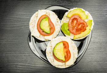A black plate with prepared healthy and dietary sandwiches, a rice cake with cheese, tomato and avocado.