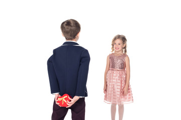 back view of boy in fashionable suit holding heart shaped gift box and looking at beautiful little girl in pink dress isolated on white