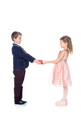 side view of stylish boy presenting heart shaped gift box to beautiful little girl isolated on white