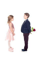 side view of stylish boy in suit holding rose flower and looking at beautiful little girl in pink dress isolated on white
