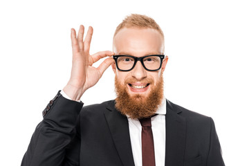 bearded young businessman adjusting eyeglasses and smiling at camera isolated on white