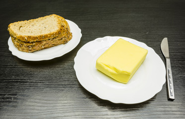 Butter and bread on white plates and a knife on the table.